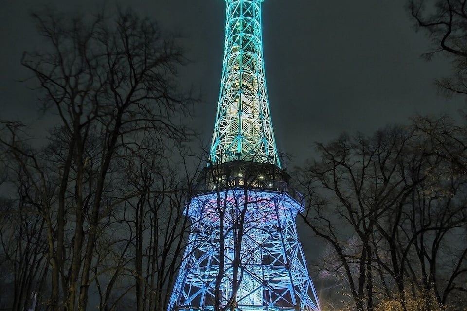 The Eiffel Tower at night, illuminated with blue lights, surrounded by trees and a park area.