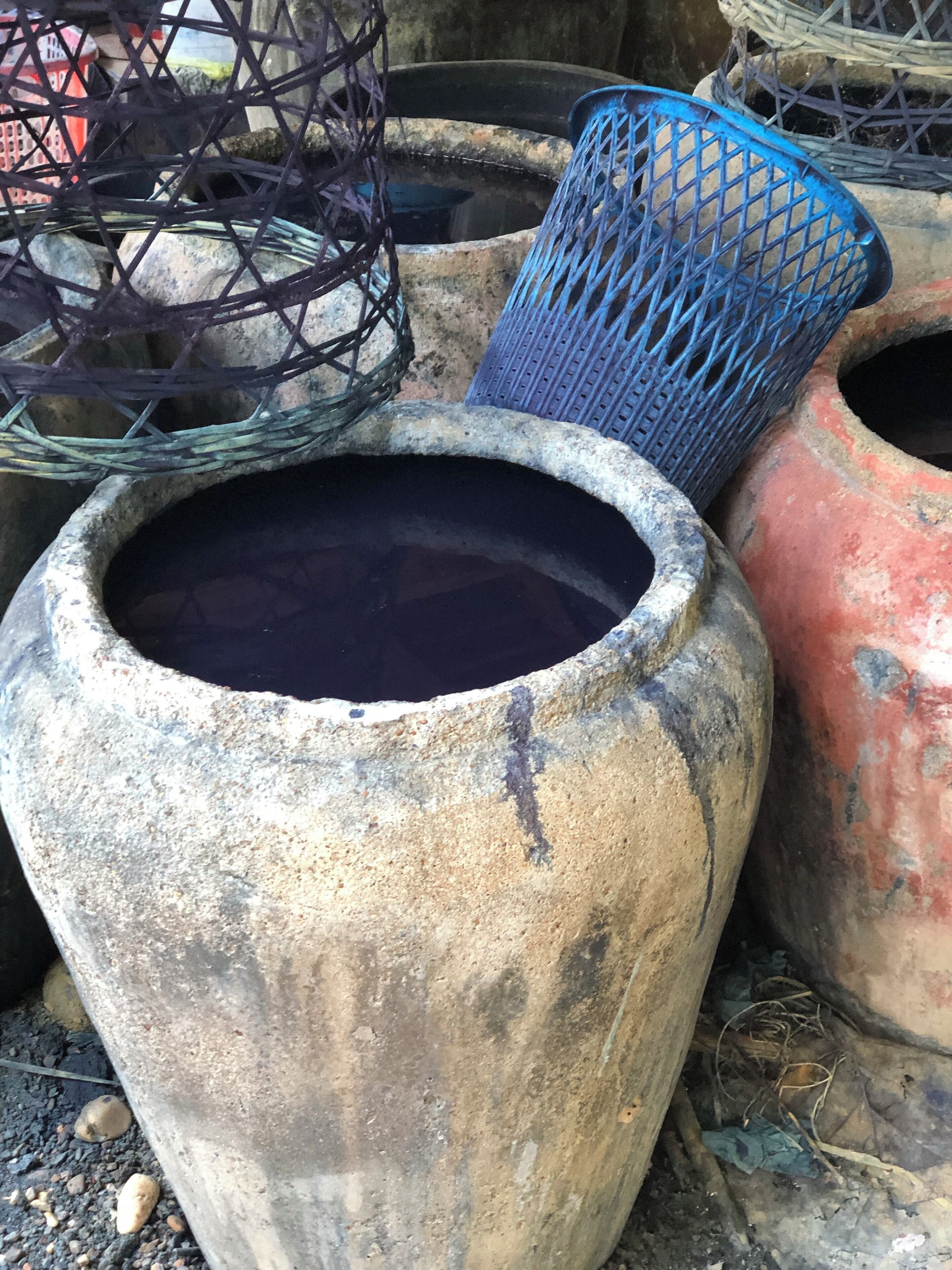 A large, weathered clay pot with a dark liquid inside, surrounded by other pottery and baskets.
