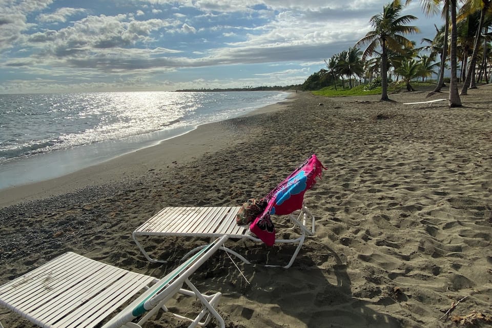 A beach with two lounge chairs and a colorful umbrella, palm trees, and a cloudy sky.