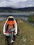 Una pequeña embarcación con motor en la orilla cubierta de hierba de un lago, con montañas y nubes al fondo.