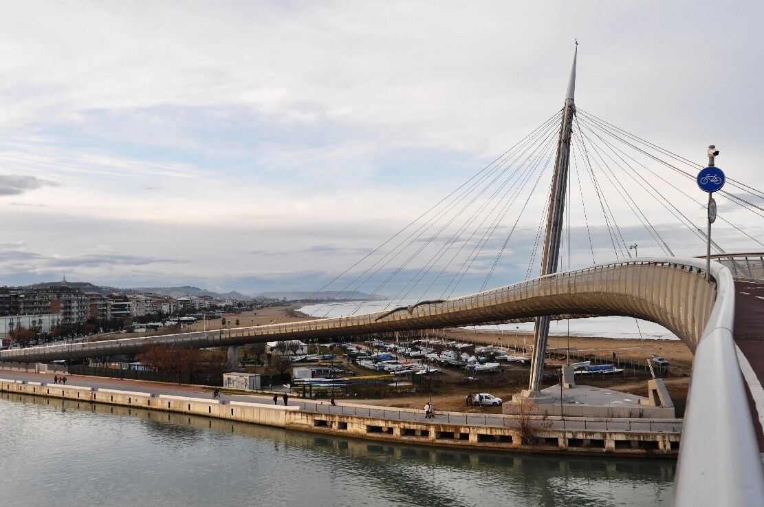 A cable-stayed bridge with a curved walkway over a body of water, with buildings and boats in the background.
