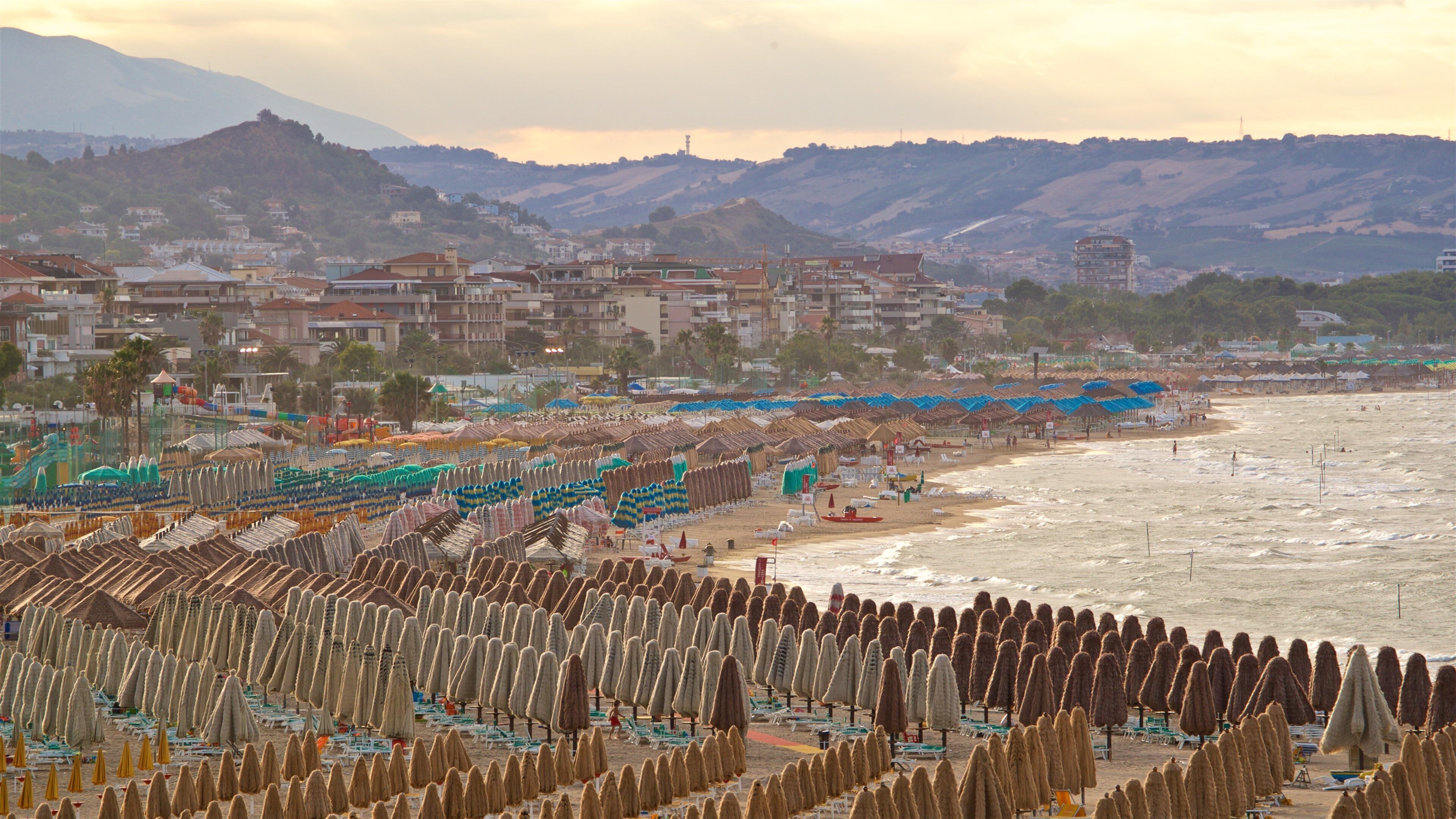 Una playa con hileras de sombrillas cónicas y sillas de playa, una orilla arenosa y edificios al fondo.