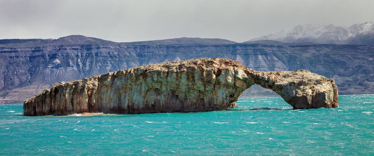 A natural rock arch formation in turquoise waters with a mountainous backdrop.