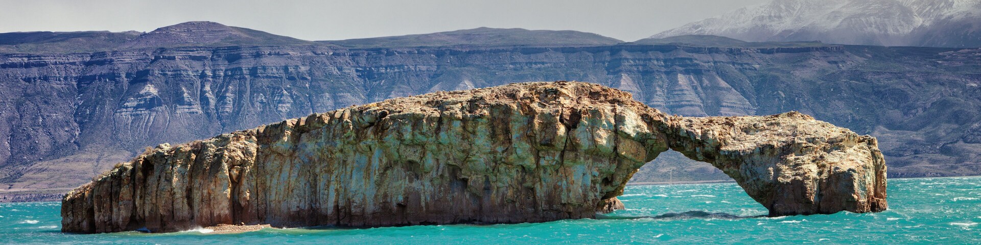 A natural rock arch formation in turquoise waters with a mountainous backdrop.
