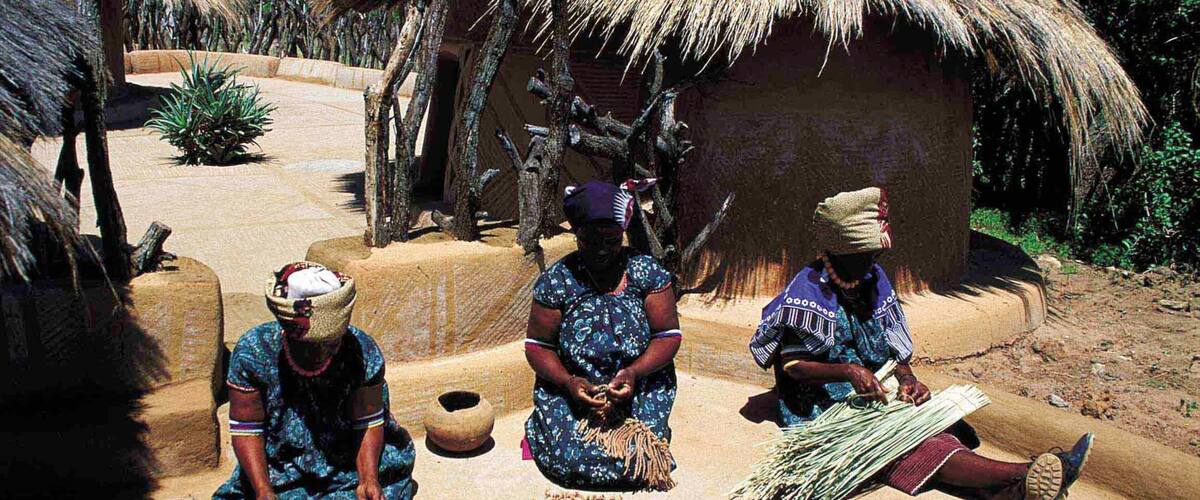 Three women in traditional attire sitting outdoors near thatched huts, with pottery and woven materials around them.