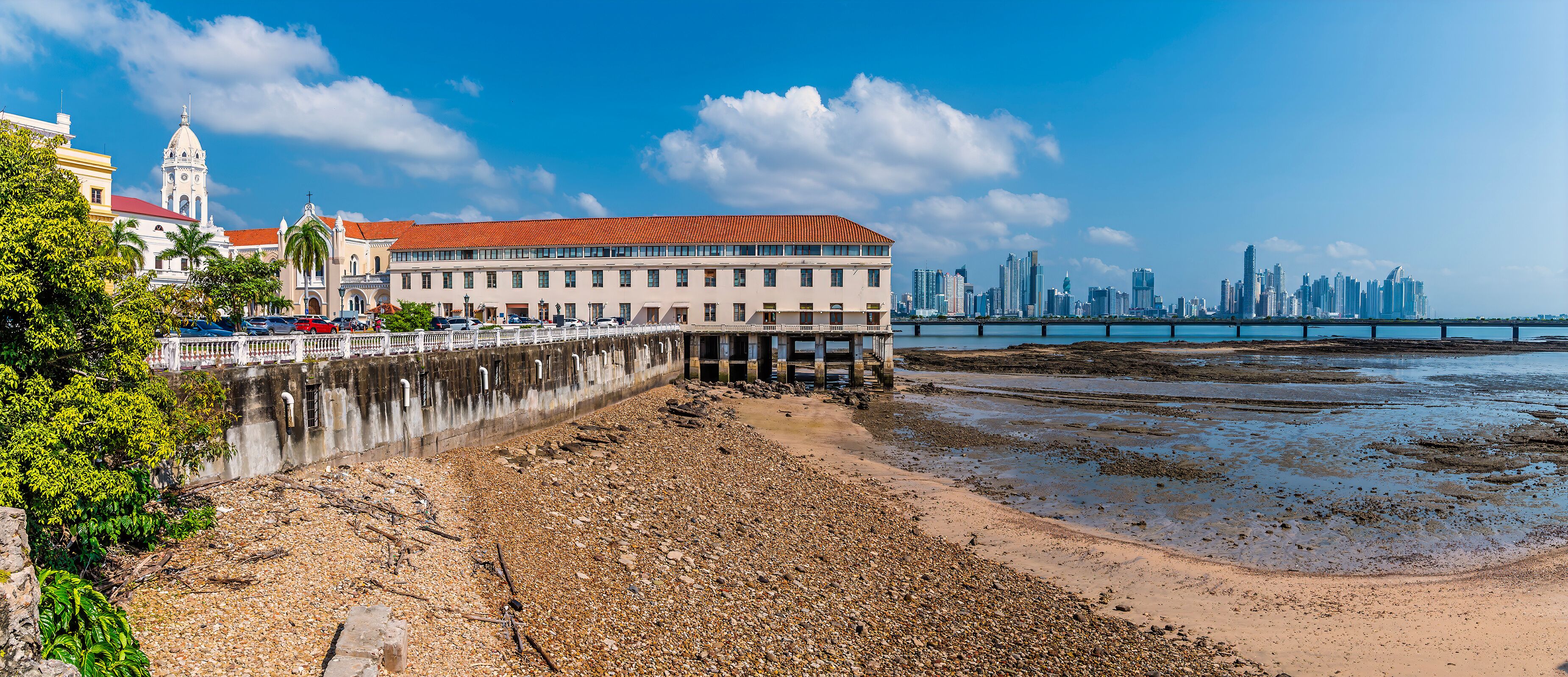 A coastal cityscape with a prominent white building with a red roof, a historic church, and a modern skyline in the background.