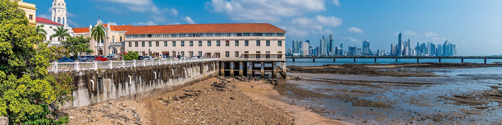 Un paisaje urbano costero con un edificio blanco destacado con techo rojo, una iglesia histórica y un horizonte moderno al fondo.