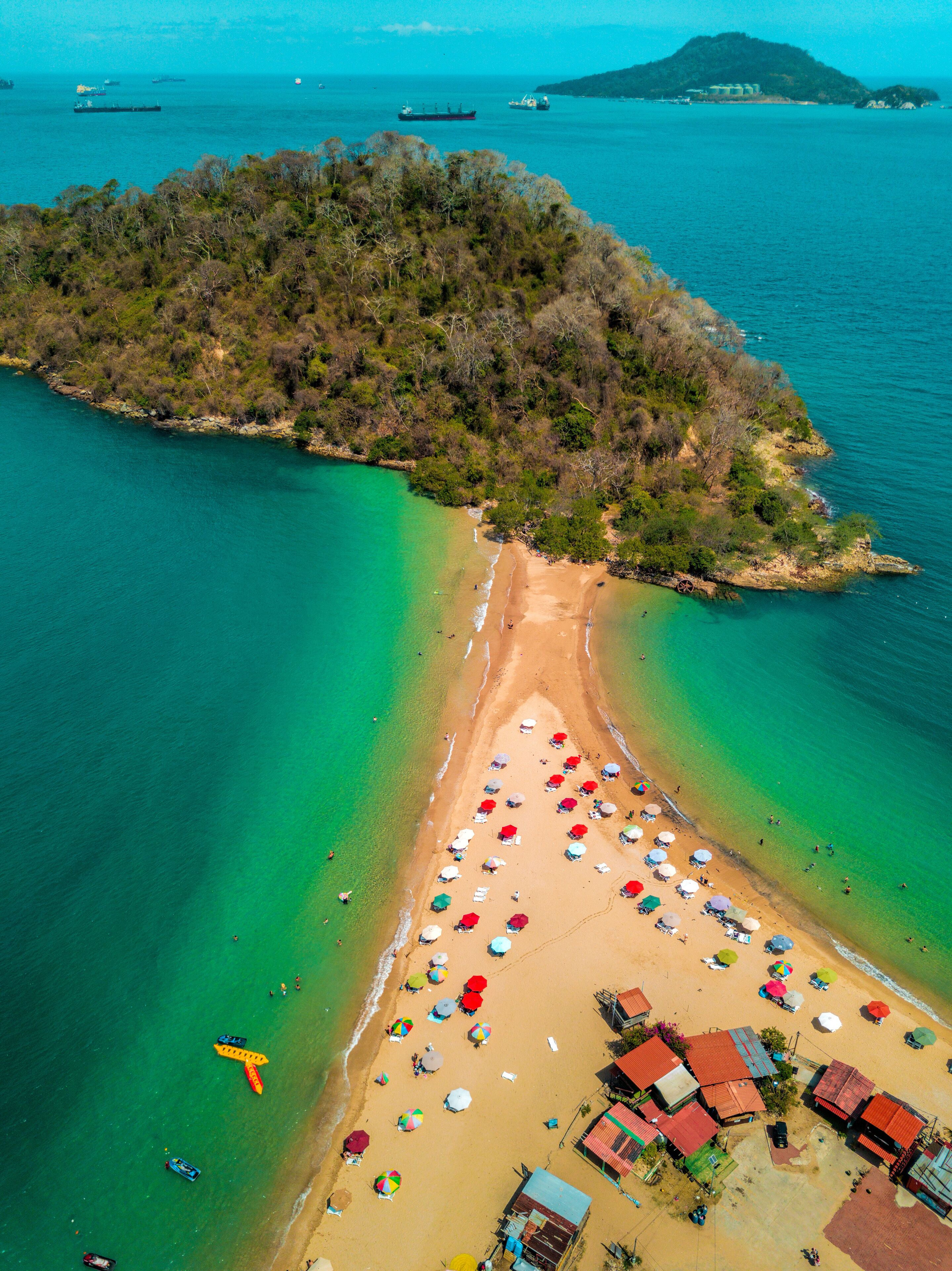 An aerial view of a tropical island with a beach, clear turquoise waters, and a small settlement with colorful umbrellas.