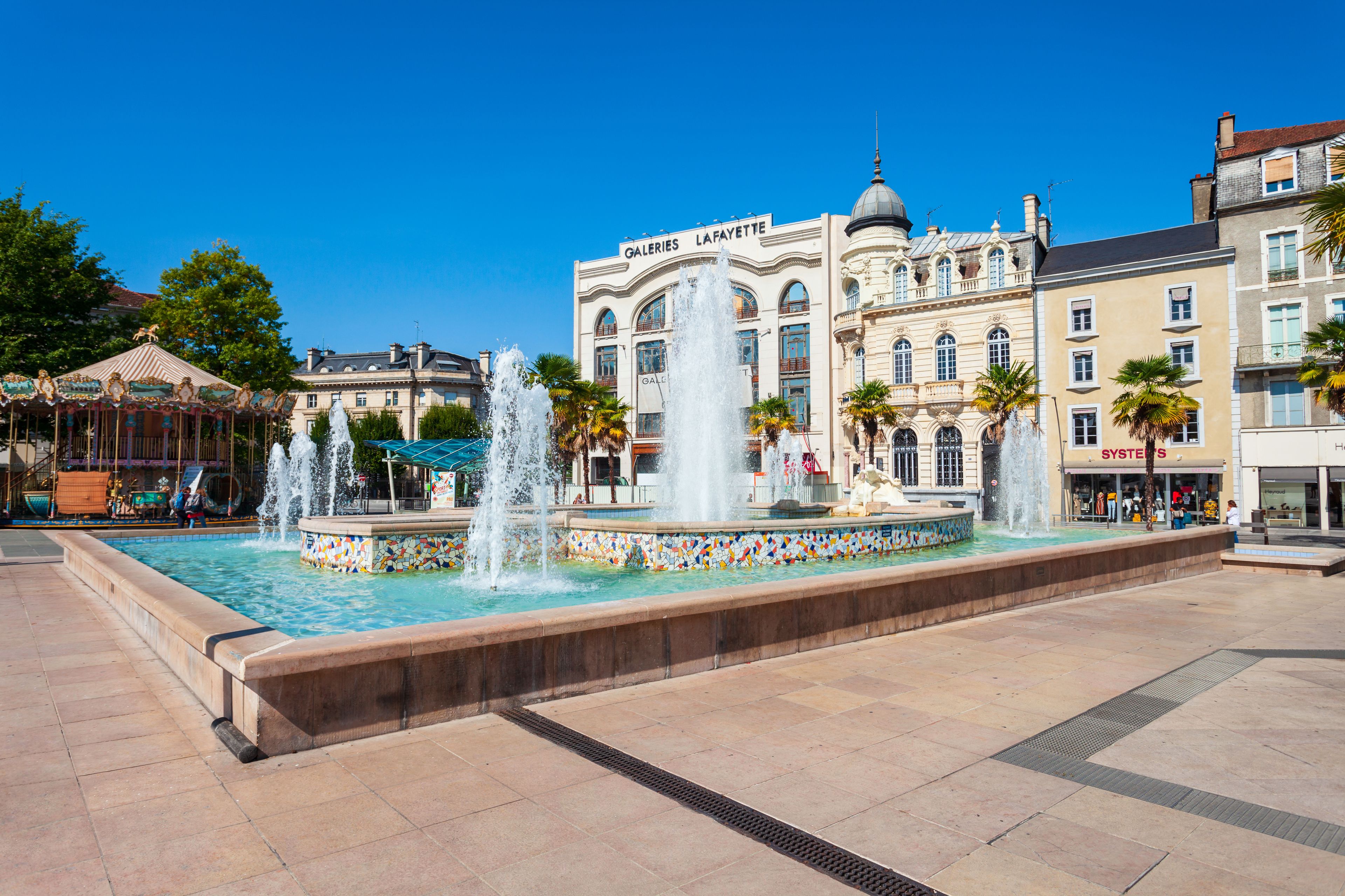A public square with a central fountain, surrounded by historic buildings and palm trees.