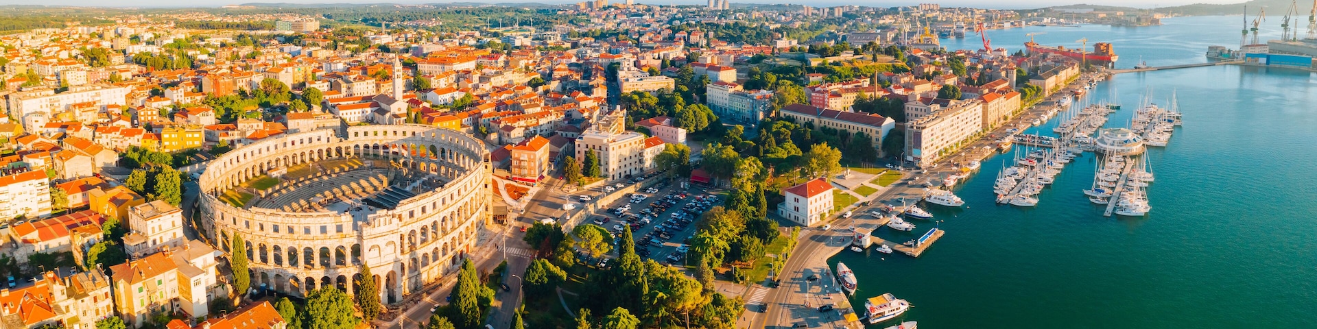 An aerial view of a coastal city with a historic amphitheater, a marina with boats, and a bridge in the distance.
