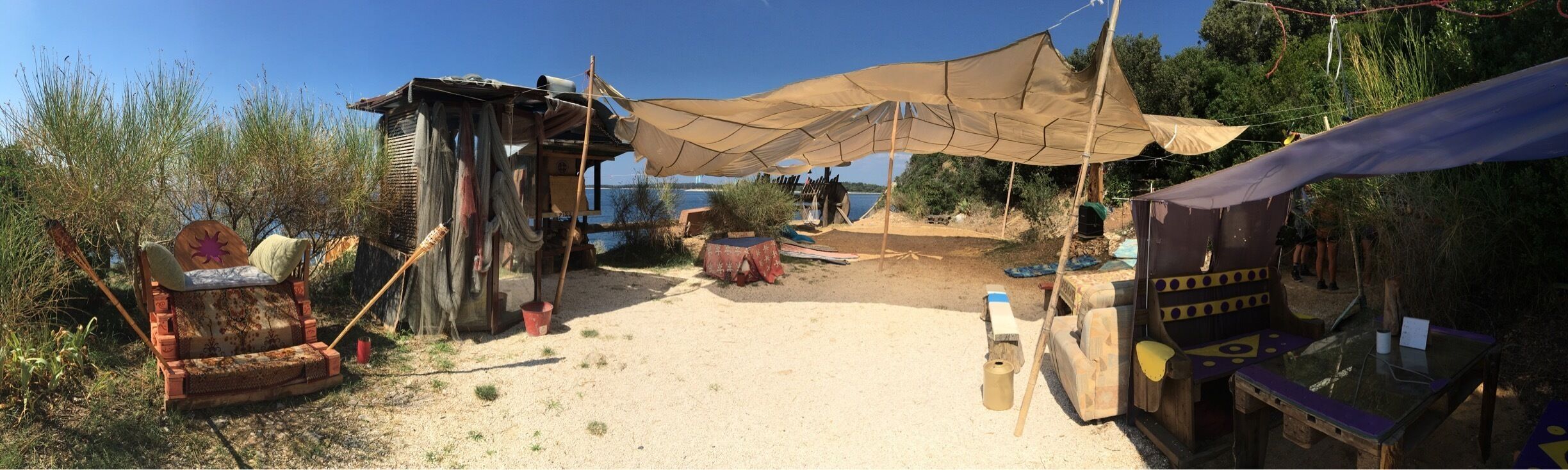 A rustic outdoor setting with a sandy ground, a wooden structure, a large canopy, and a small table with items on it.