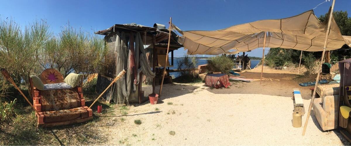A rustic outdoor setting with a sandy ground, a wooden structure, a large canopy, and a small table with items on it.