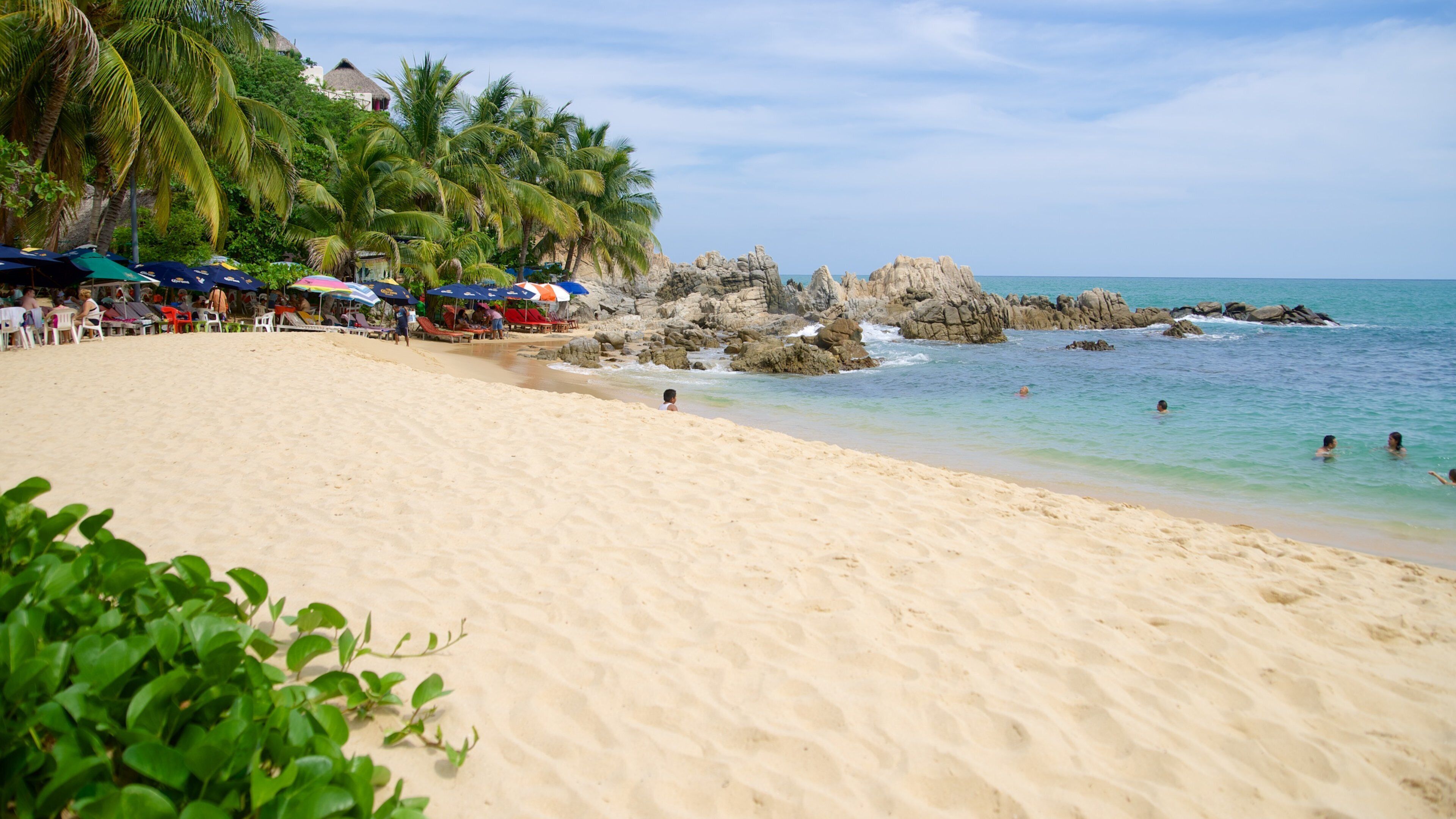 A sandy beach with palm trees, a rocky outcrop, and people swimming.