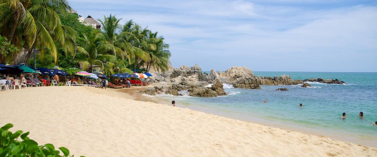 A sandy beach with palm trees, a rocky outcrop, and people swimming.