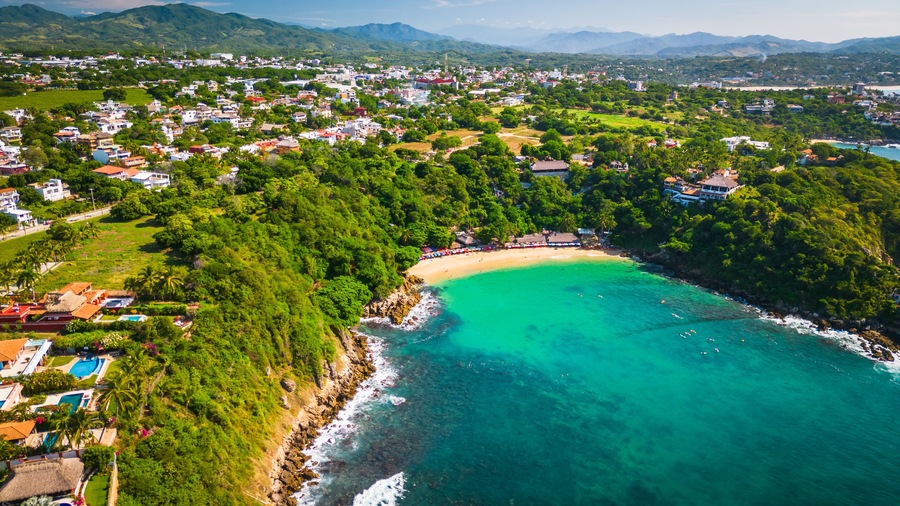 Un pueblo costero con playa, abundante vegetación y montañas al fondo.