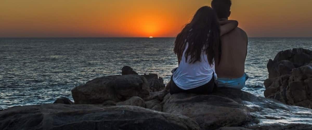 A couple sitting on rocks by the sea during sunset.