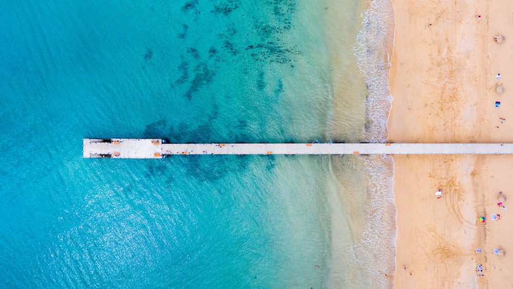 A pier extending into turquoise waters with a sandy beach and scattered beachgoers.