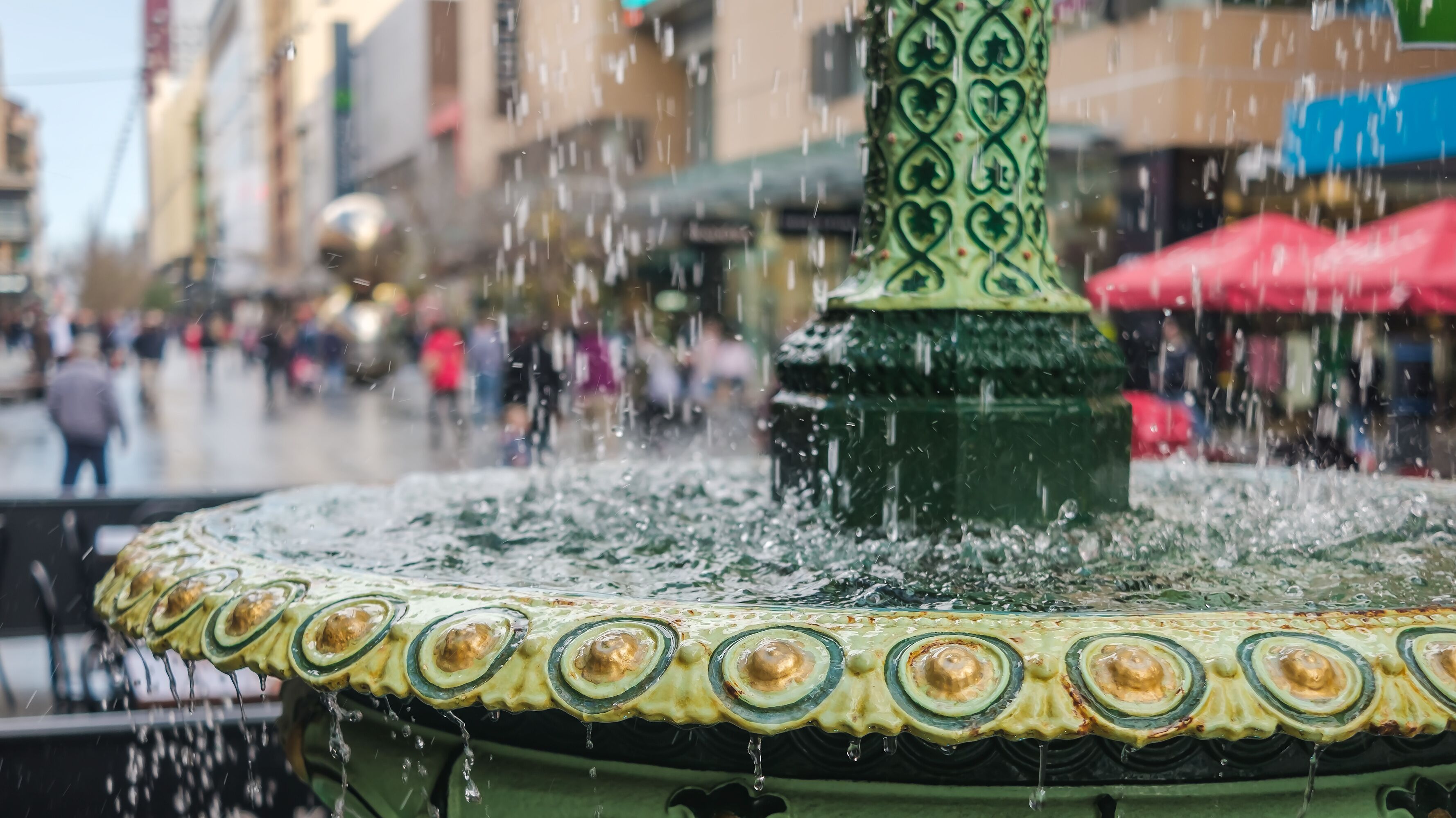 Uma fonte com coluna verde e anéis decorativos, em uma praça urbana movimentada com figuras borradas e edifícios.