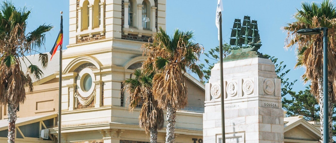 Um edifício histórico com uma torre do relógio, palmeiras e uma estátua em uma praça pública.