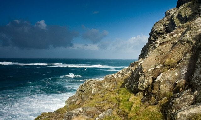 A rocky coastline with waves crashing against the shore under a partly cloudy sky.