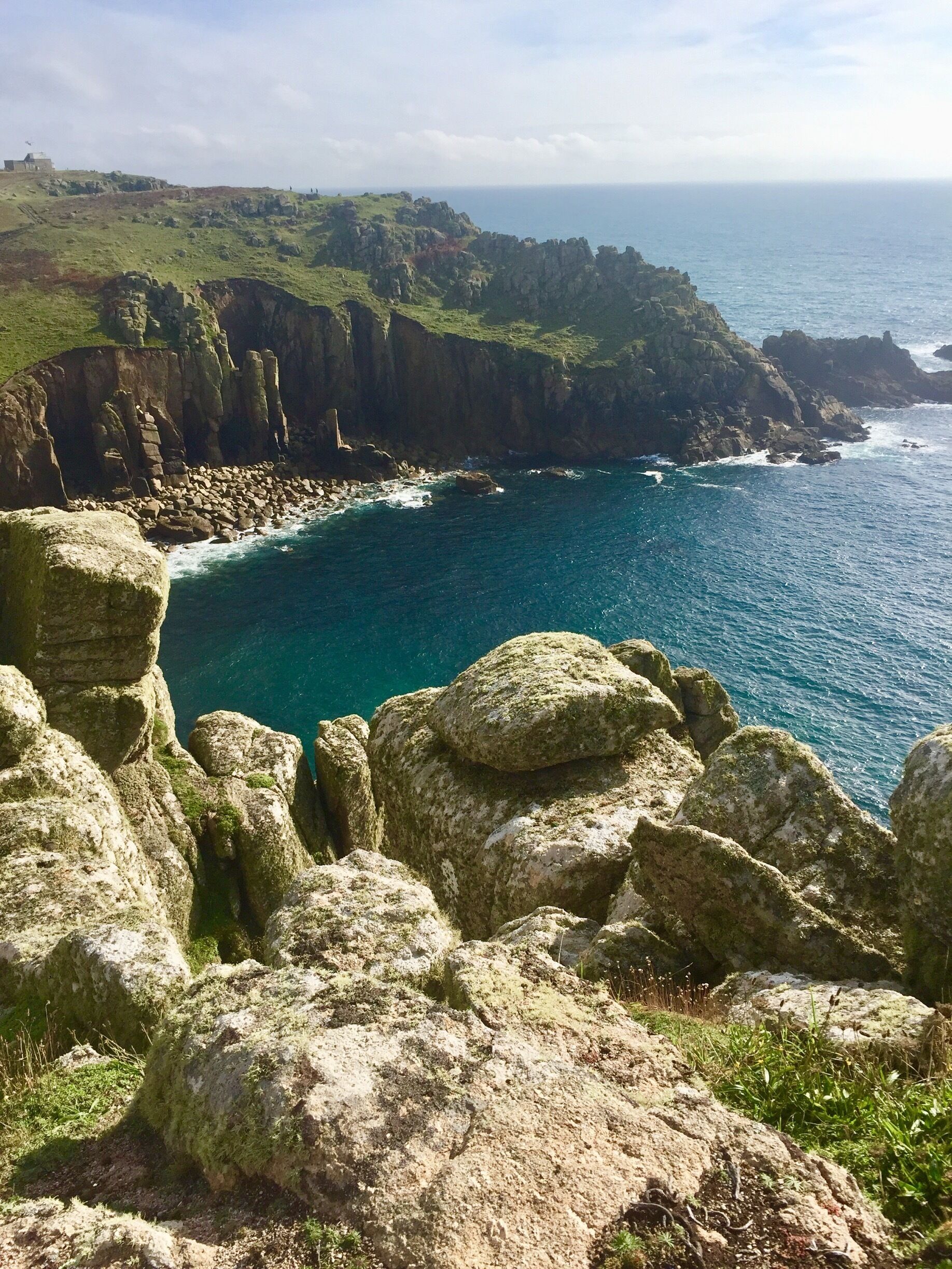A coastal landscape with rocky cliffs, a clear blue sea, and a grassy hill.