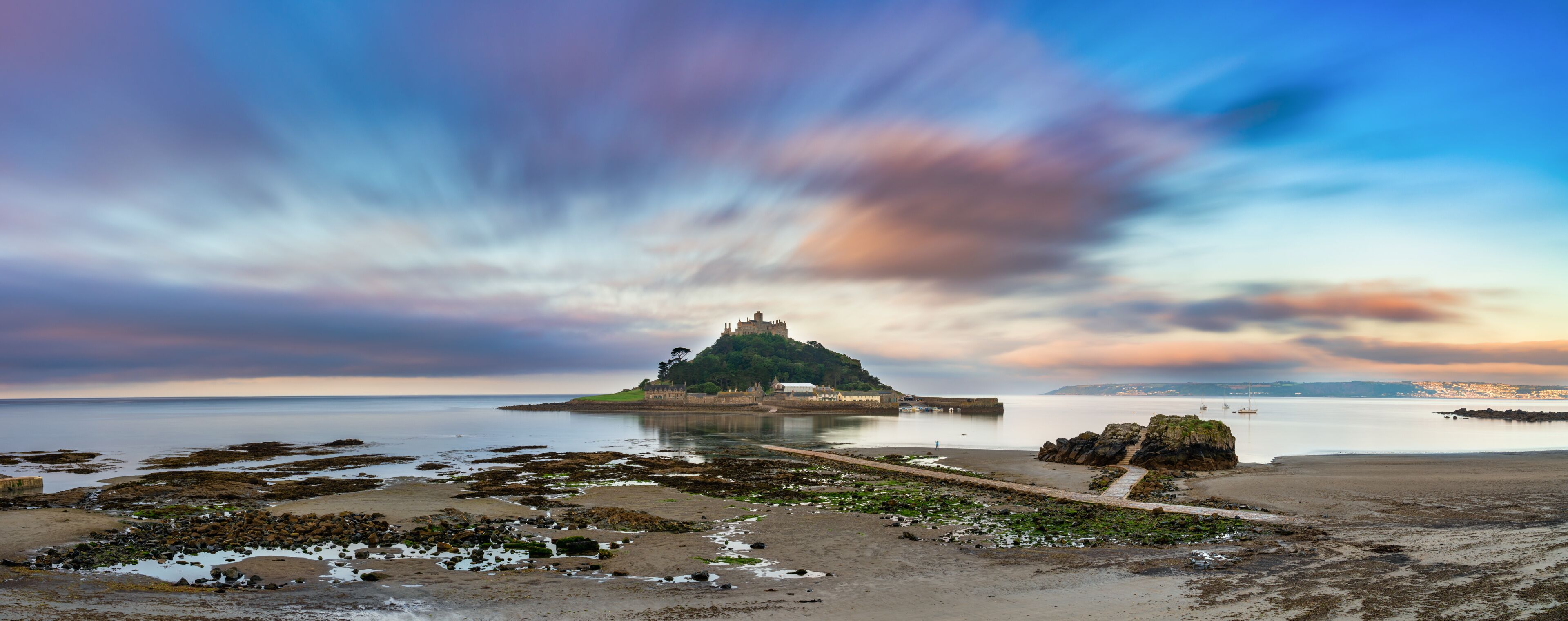 A coastal landscape with a castle on a small island, calm water, and a rocky shoreline.