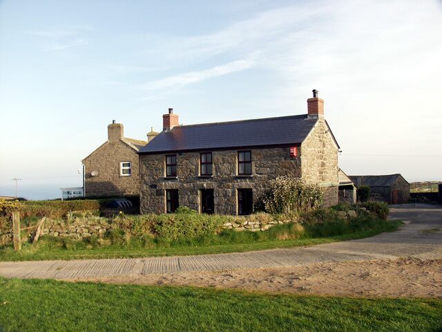 A stone house with a sloped roof and chimneys, surrounded by a grassy area and a paved path.