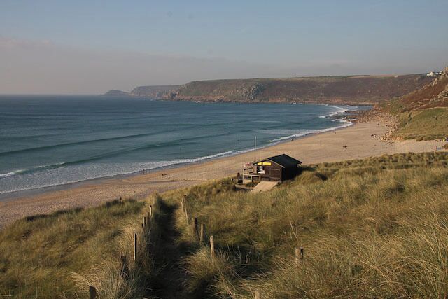 A beach with a small house on the dunes, the ocean, and a distant cliff.