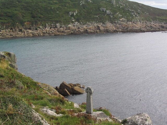 Paisagem costeira com um monumento de pedra, litoral rochoso e colinas verdes ao fundo.