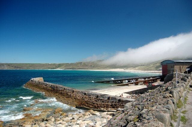 A rocky shoreline with a breakwater, calm sea, and a building in the distance.