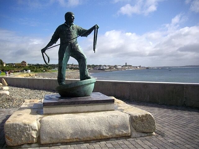 Una estatua de un hombre sosteniendo una caña de pescar sobre un pedestal, junto a la orilla del mar.
