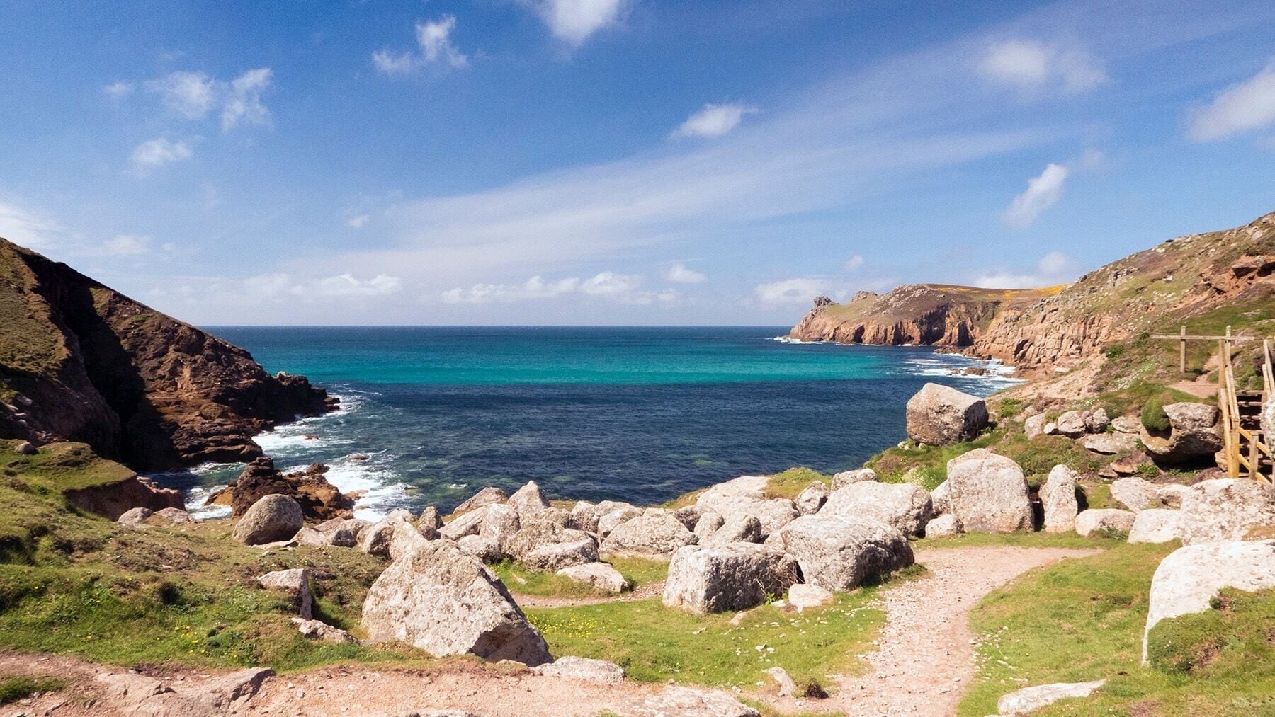 A coastal landscape with rocky terrain, a clear blue sea, and a wooden pathway.