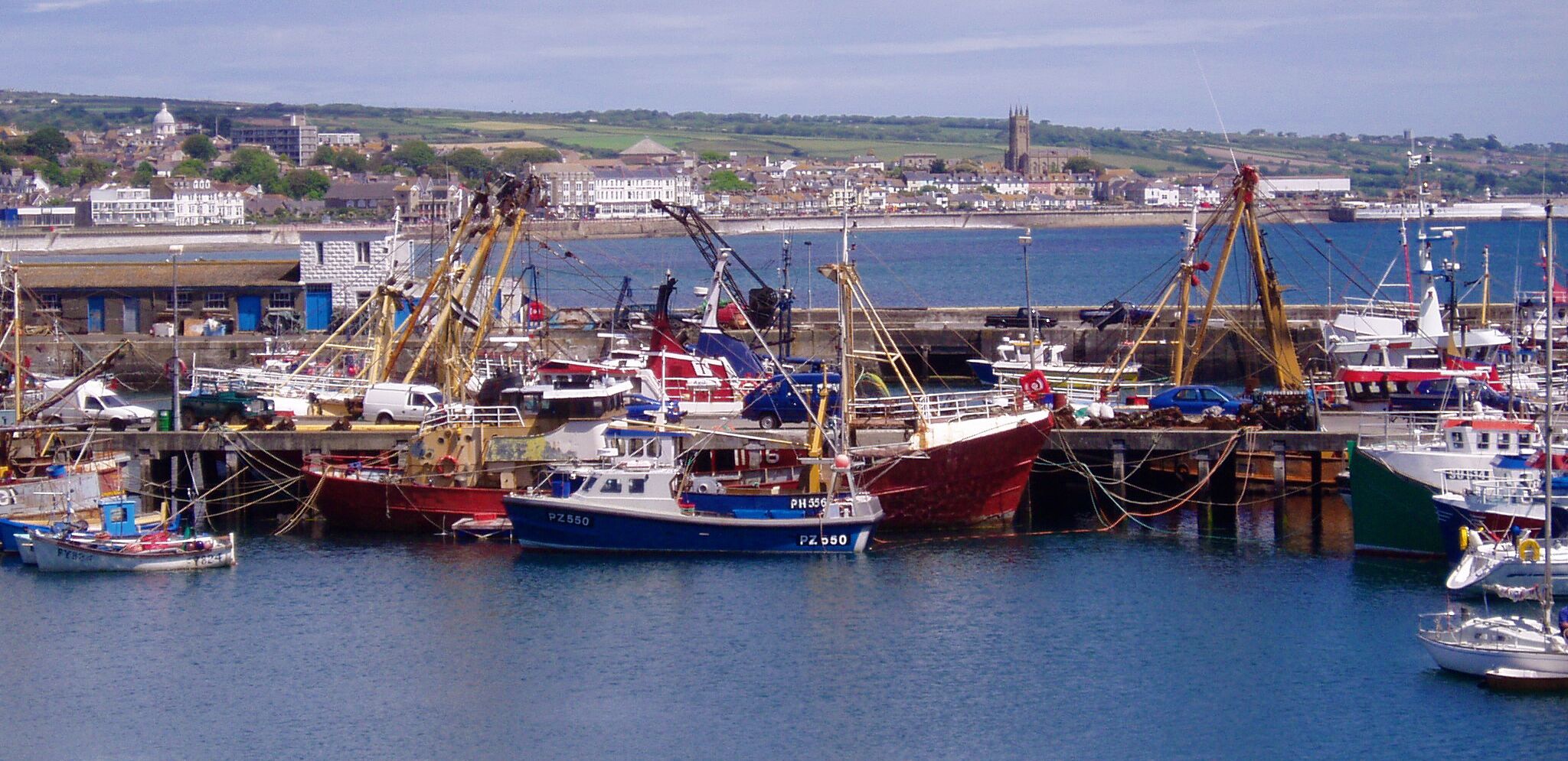 A harbor with several fishing boats docked, a crane, and a distant church.