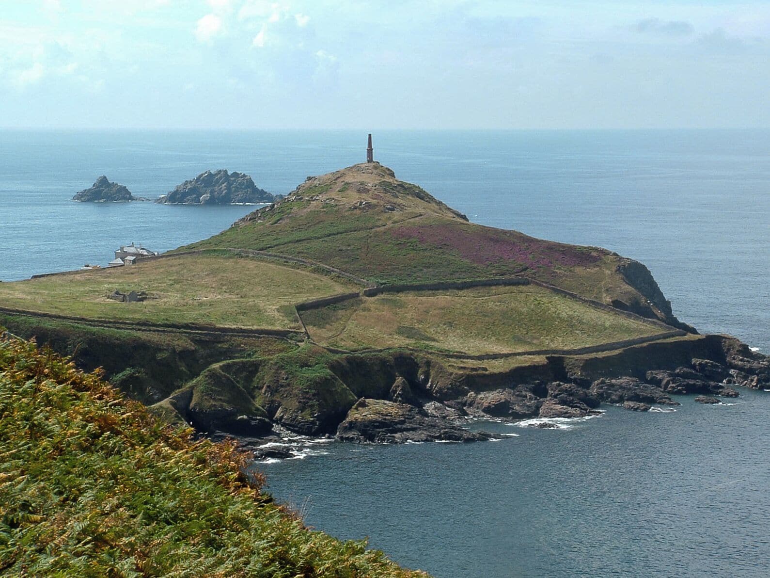 A coastal landscape with a prominent lighthouse on a hill, rocky outcrops, and a grassy area with purple vegetation.
