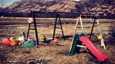 A playground with swings and a slide, surrounded by a grassy field and mountains in the background.