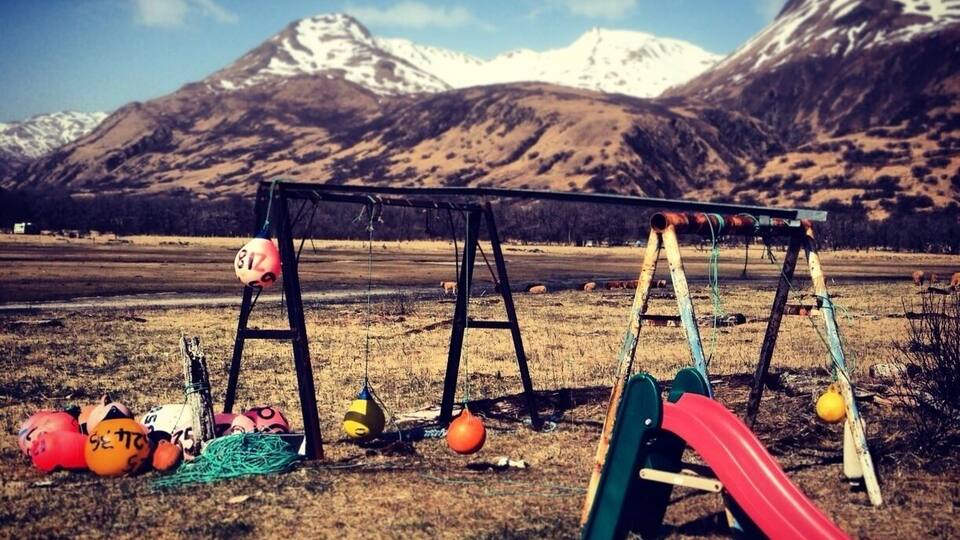A playground with swings and a slide, surrounded by a grassy field and mountains in the background.