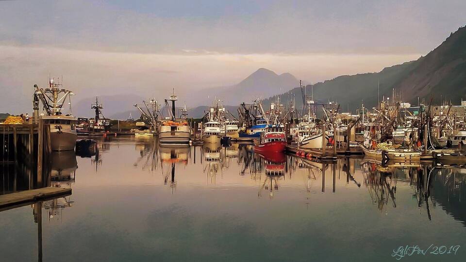 A harbor with numerous boats docked, calm water, and a mountain in the background.