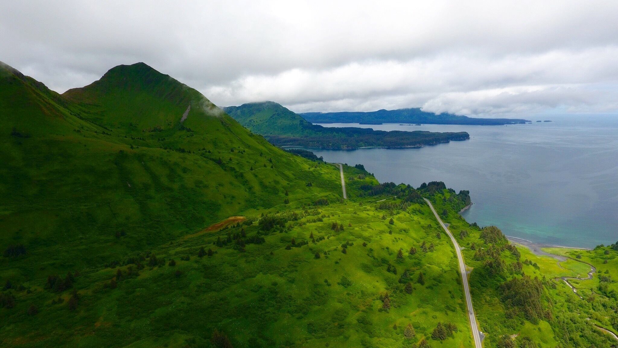 A lush green landscape with a winding road, a body of water, and distant mountains under a cloudy sky.