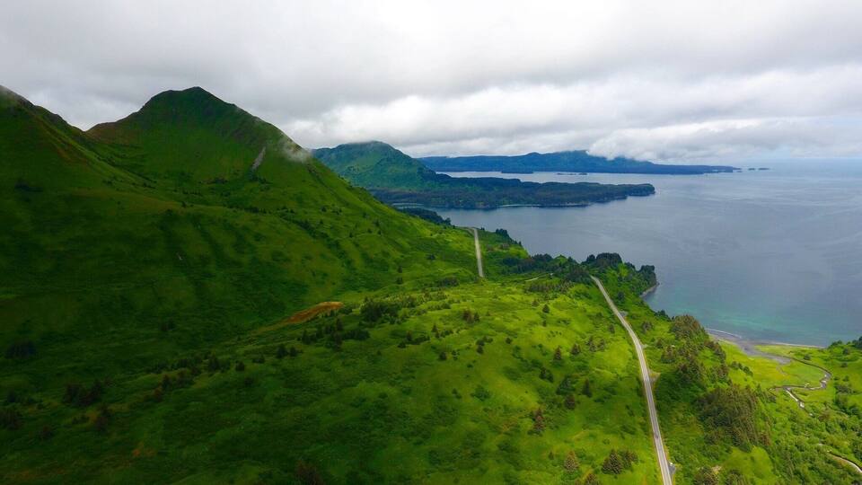 A lush green landscape with a winding road, a body of water, and distant mountains under a cloudy sky.