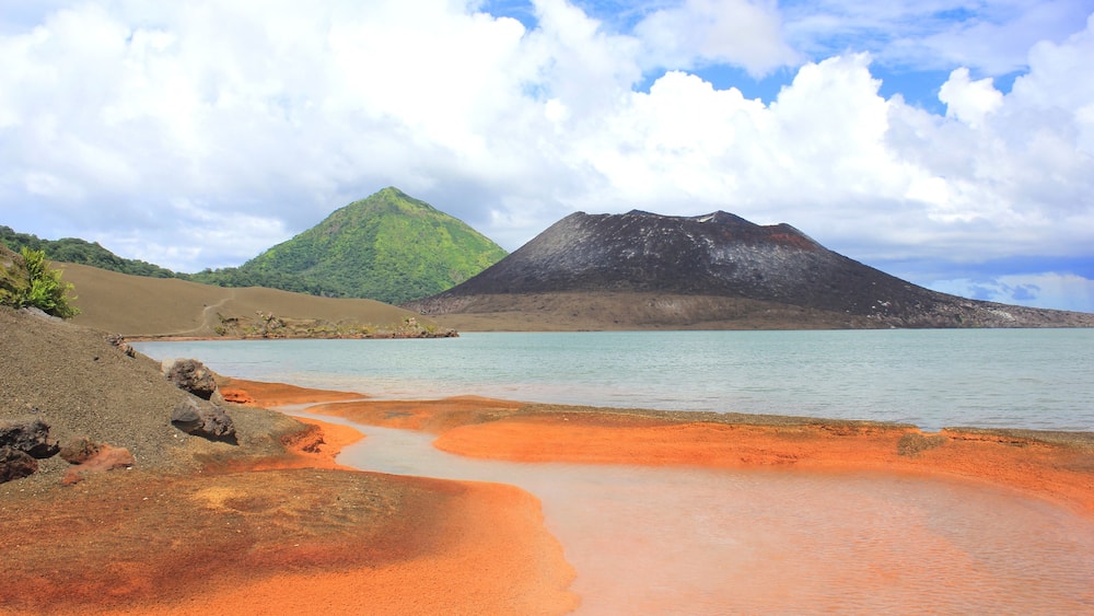 A beach with reddish sand, calm water, and two mountains in the background.