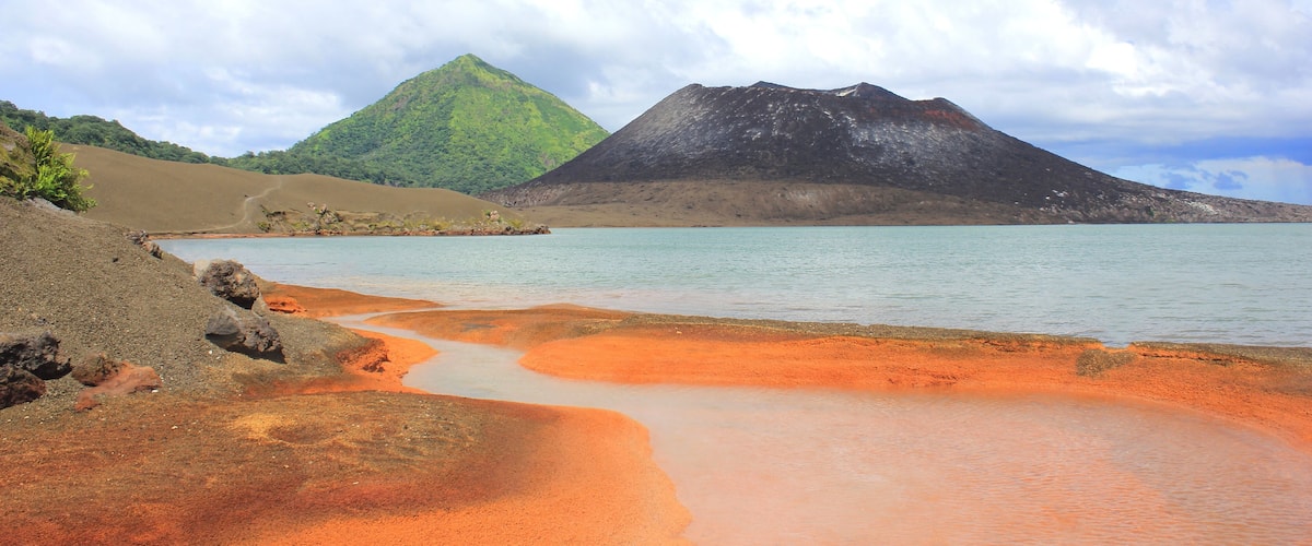 A beach with reddish sand, calm water, and two mountains in the background.