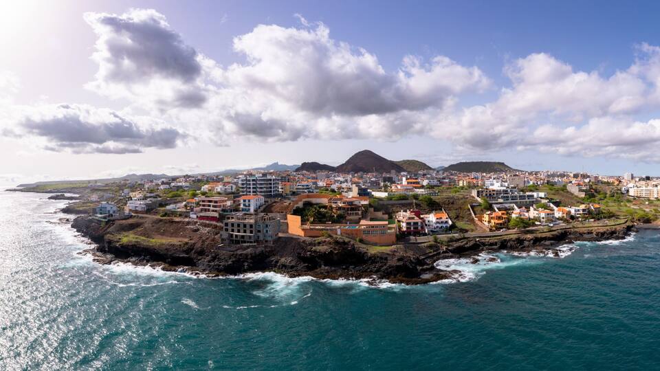 A coastal town with buildings along the shoreline, a prominent hill in the background, and the ocean in the foreground.