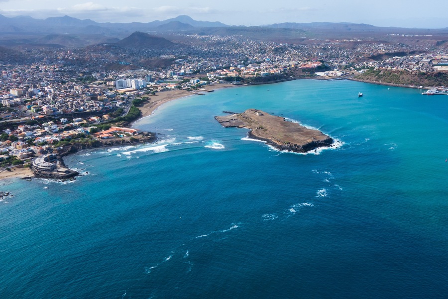 Vista aérea de una ciudad costera con una península destacada, una gran masa de agua y montañas a lo lejos.