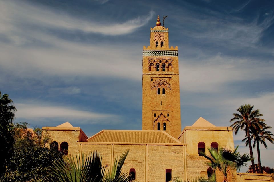 A tall, ornate tower with intricate designs and a pointed roof, surrounded by palm trees and a clear sky.