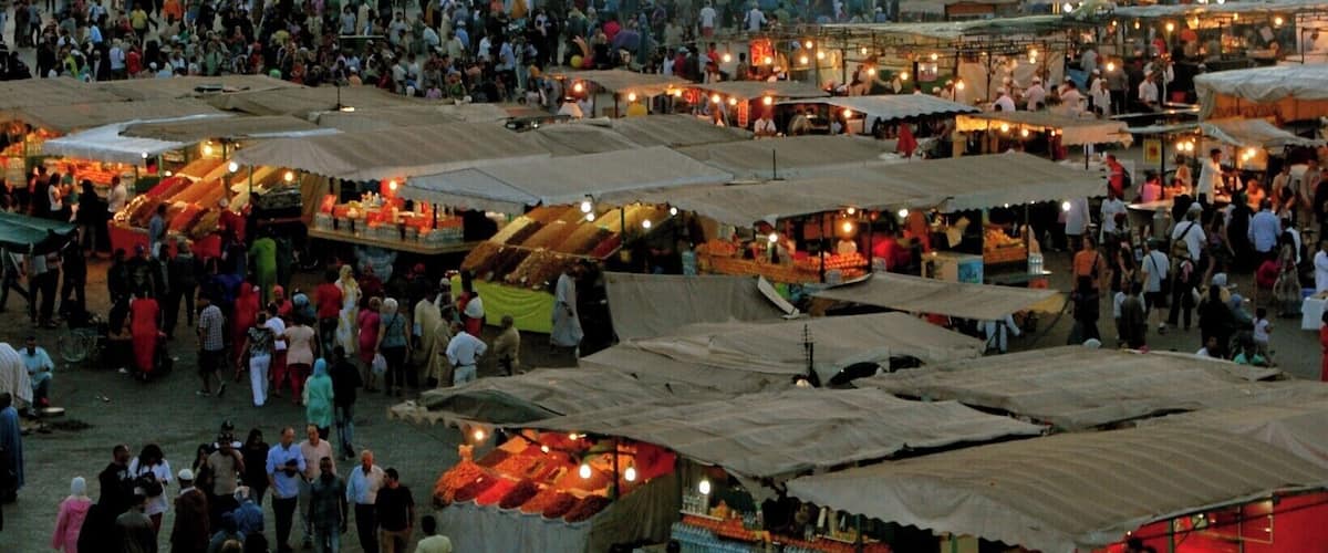A bustling outdoor market with numerous stalls and a large crowd. In the background, a tall tower with a clock is visible against a sunset sky.