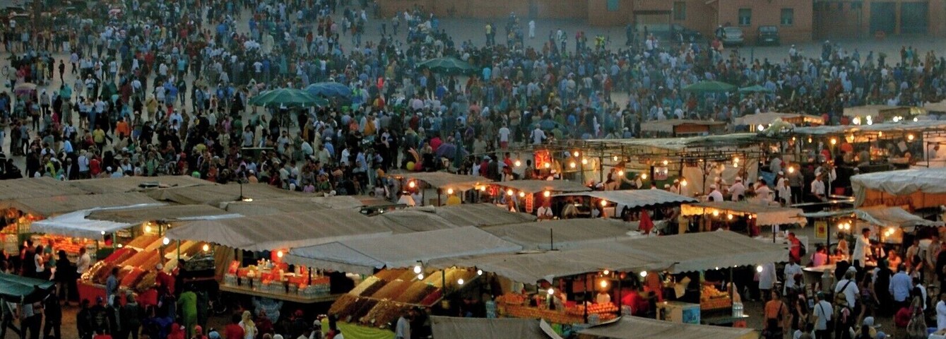 A bustling outdoor market with numerous stalls and a large crowd. In the background, a tall tower with a clock is visible against a sunset sky.