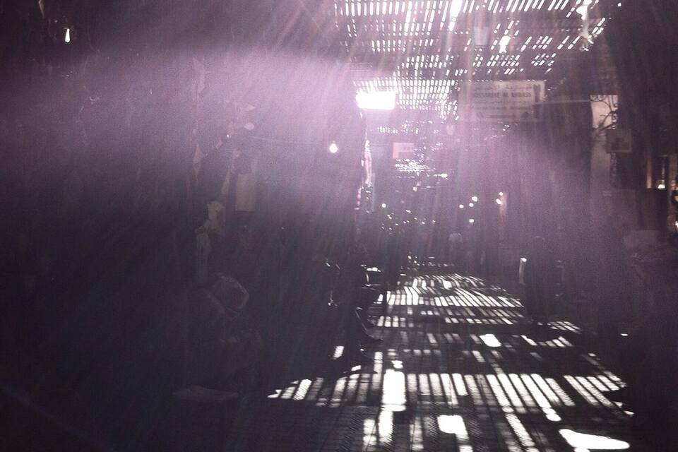 A street with a grid of shadows cast by a structure, possibly a building or a fence, under artificial lighting.