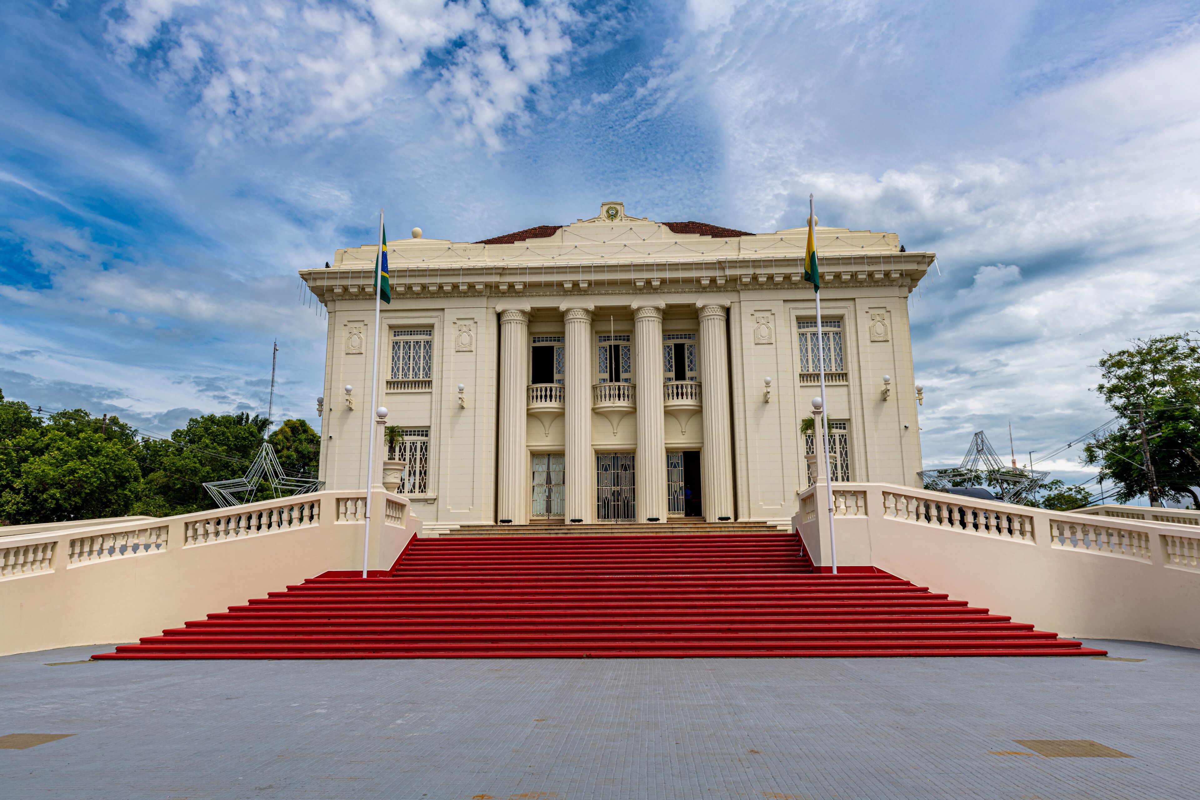 A grand building with a red staircase leading to the entrance, surrounded by a white fence and under a partly cloudy sky.