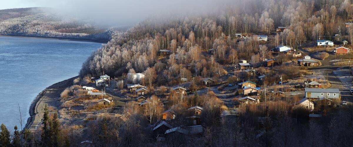 A coastal village with houses near a body of water, surrounded by forested hills.