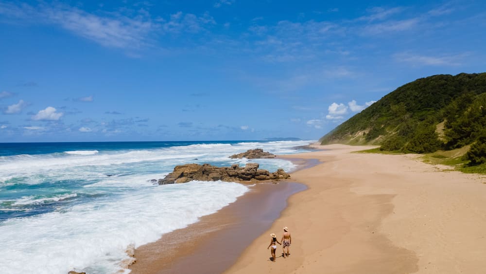Ein sandiger Strand mit Wellen, ein Paar spaziert am Strand entlang, und ein grüner Hügel ist im Hintergrund zu sehen.